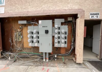 An open utility panel with multiple electrical meters mounted on a wall. Exposed wires are visible below the meters. A sign indicates the manager's apartment is #13. A laundry room is partially visible.