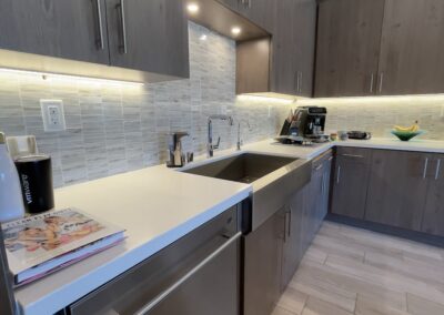 A modern kitchen featuring wooden cabinetry, a stainless steel sink, white countertops, a coffee maker, and a cookbook placed near the sink with under-cabinet lighting illuminating the backsplash.